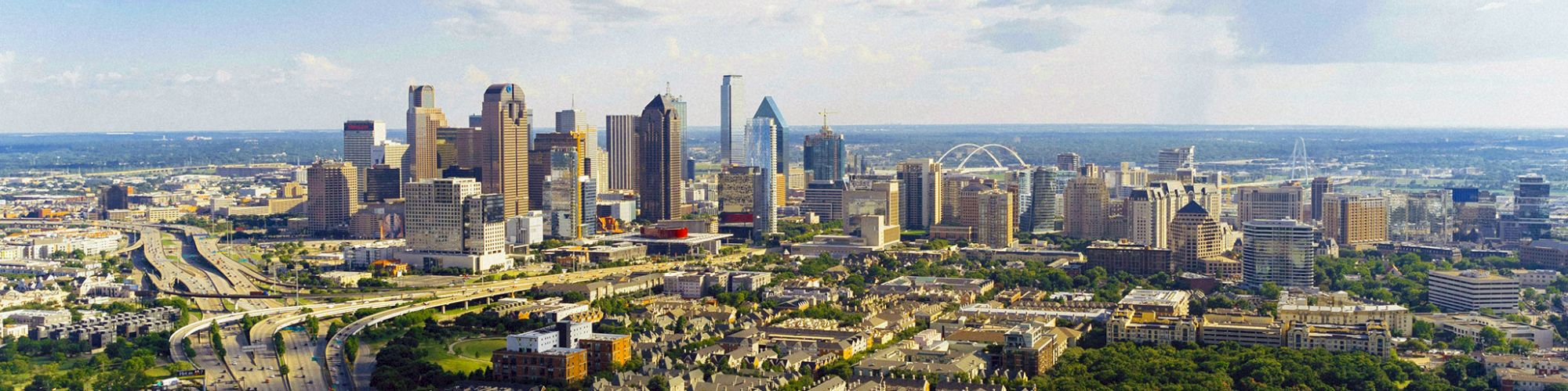 Aerial view of a city skyline with tall buildings, highways, and green spaces under a partly cloudy sky.