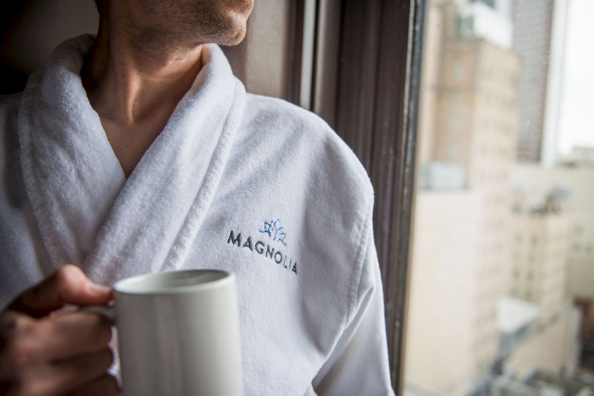 A person in a white Magnolia robe holds a mug, gazing out of a window with a blurred cityscape in the background.