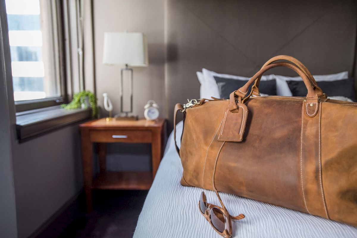 A leather duffel bag and sunglasses are on a bed in a hotel room with a bedside table, lamp, and window.