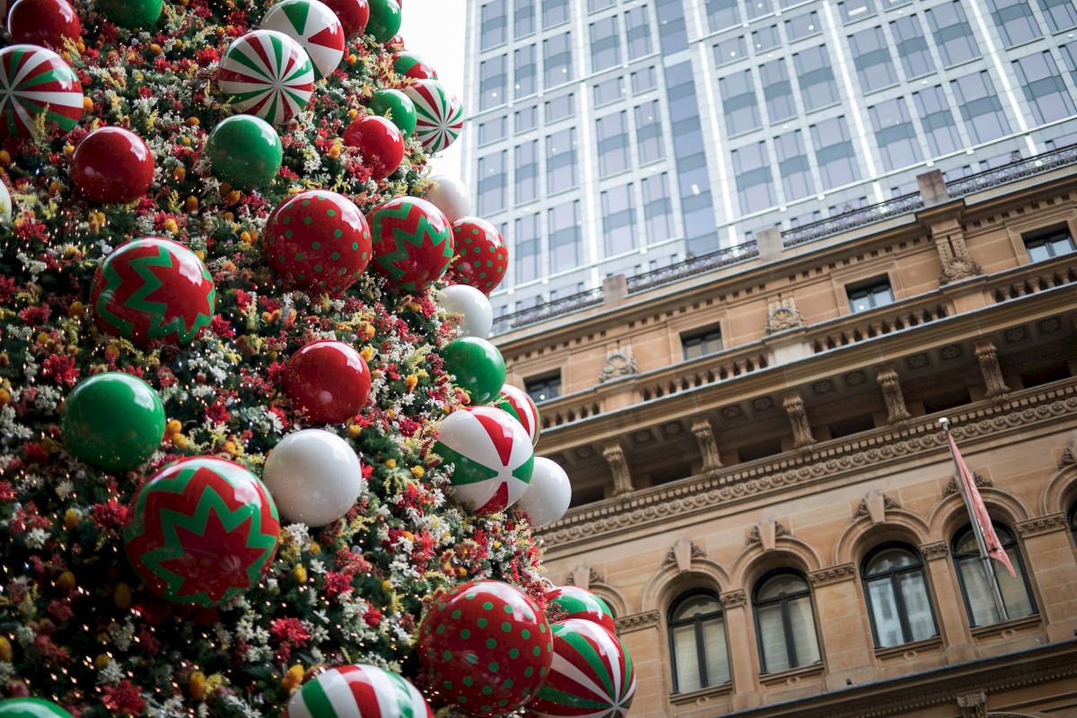 The image shows a festive Christmas tree adorned with colorful ornaments in front of a large building with classic architecture.