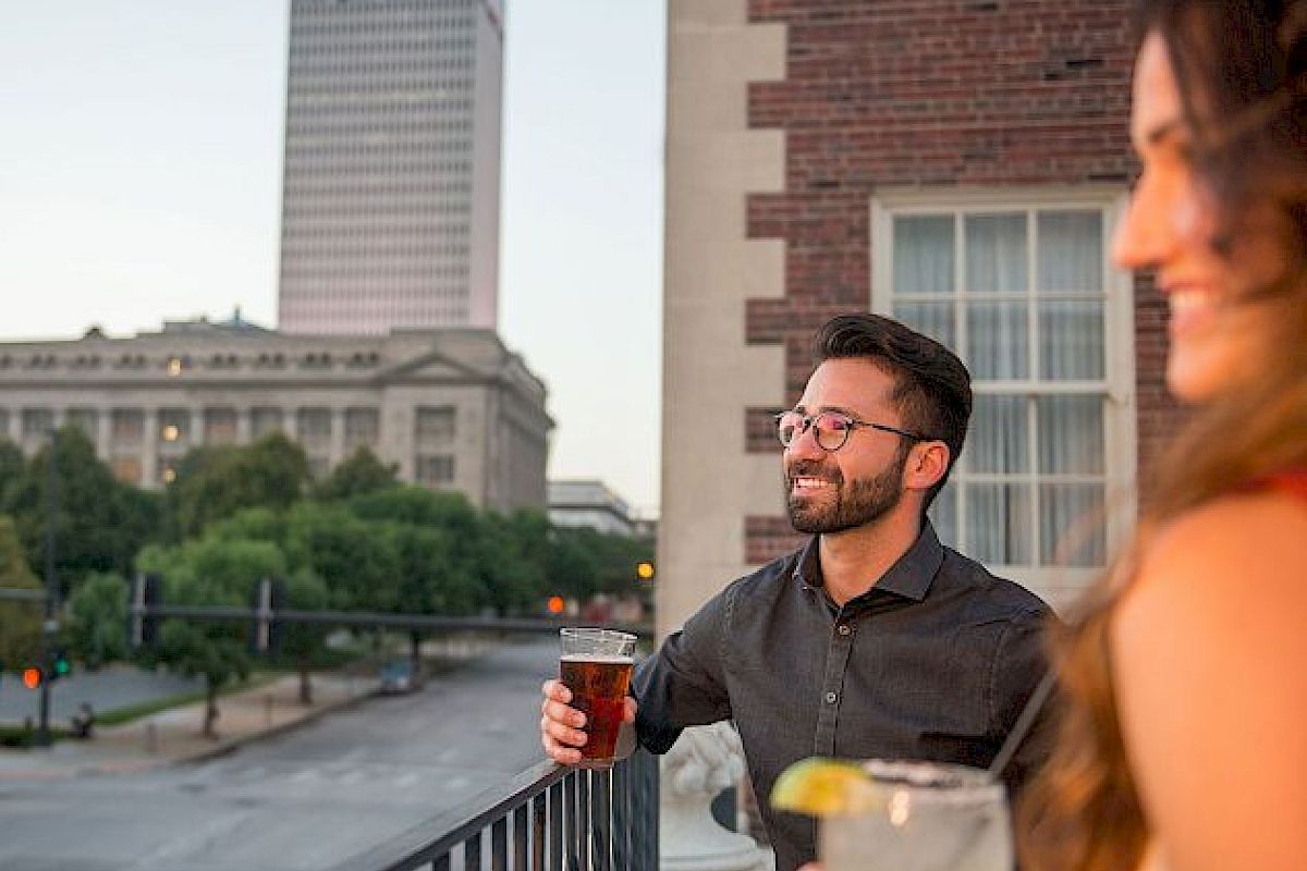A man and woman enjoy drinks on a balcony with a city view, featuring tall buildings and trees in the background.