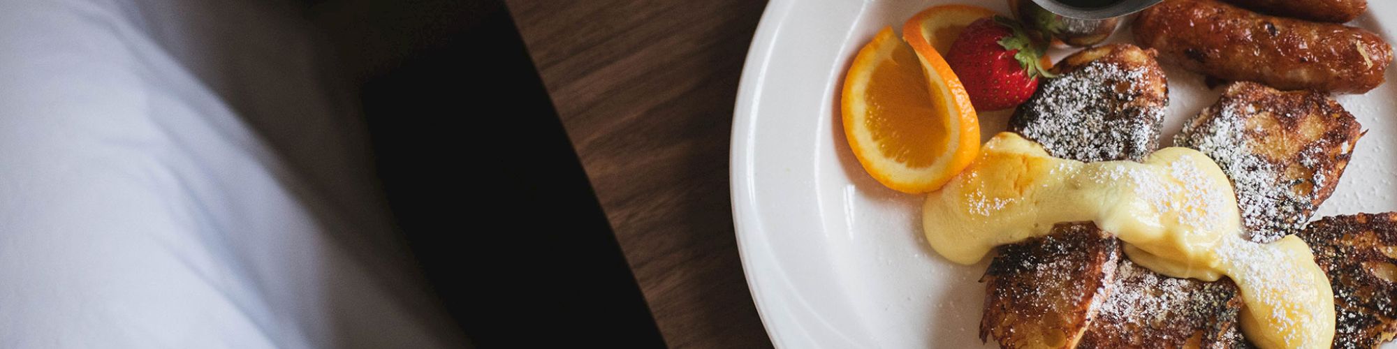 A plate of French toast with powdered sugar, syrup, orange slices, strawberries, sausages, and a cup of coffee next to a bed.