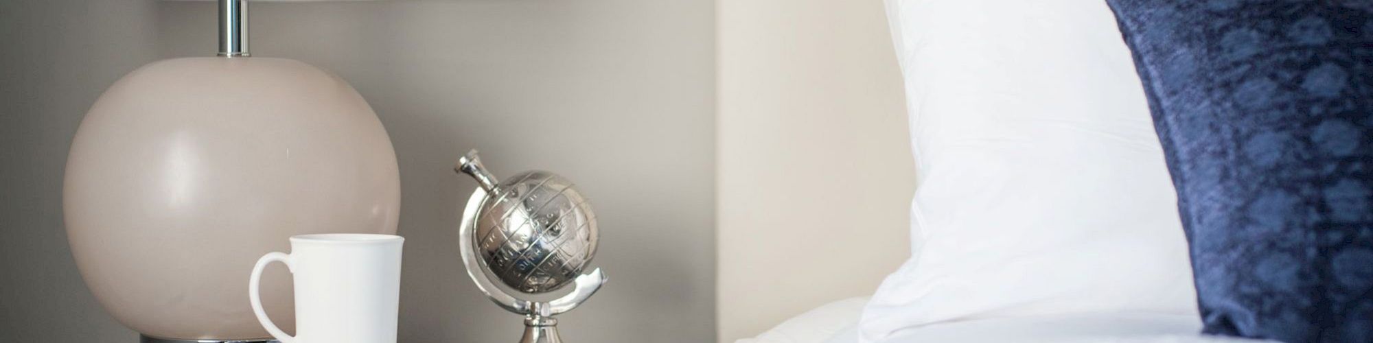 A bedside table with a lamp, a small globe, books, and a white mug next to a bed with white and blue pillows ends the sentence.