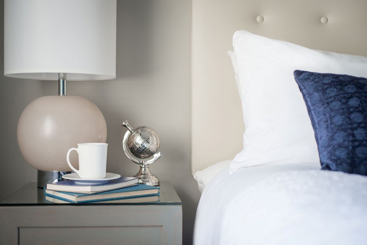 A bedside table with a lamp, books, a silver globe, and a white mug. A bed with white linens and a blue pillow is beside it.