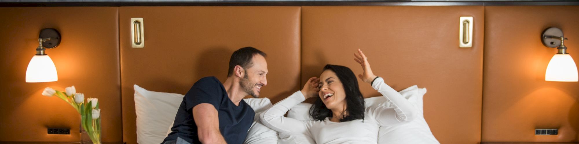 A couple relaxes on a bed with framed art on the wall, under warm lighting in a cozy room with bedside lamps and tulips.