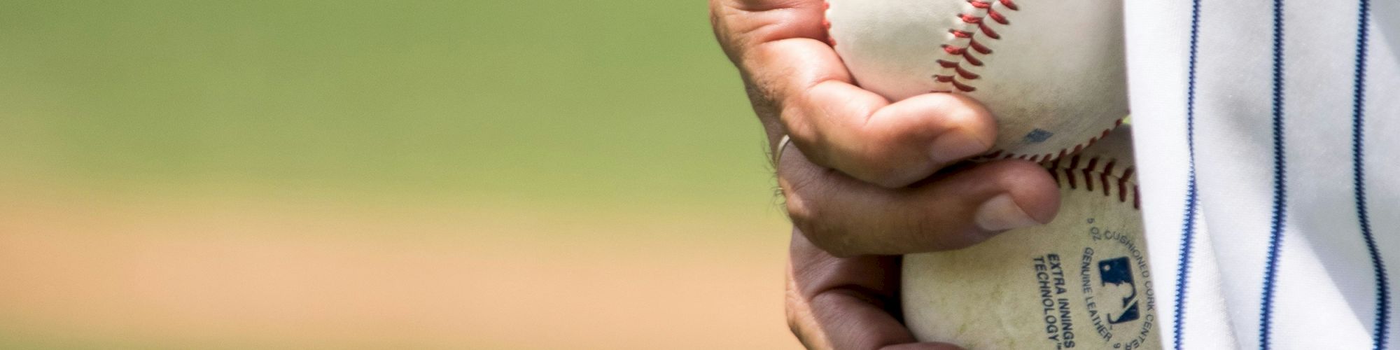 A person is holding two baseballs, wearing a pinstripe uniform, with a blurred baseball field in the background.