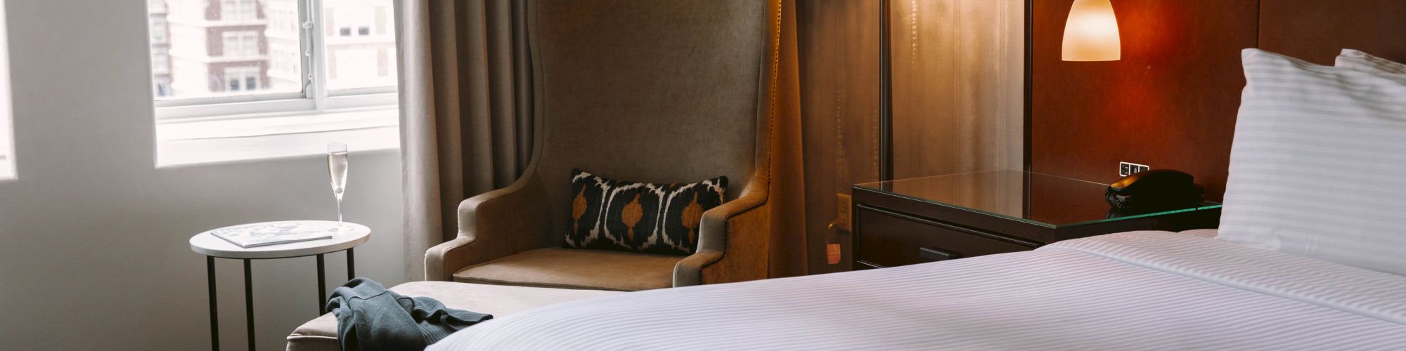 A cozy hotel room with a bed, armchair, side table, and window, decorated with warm lighting and neutral tones.