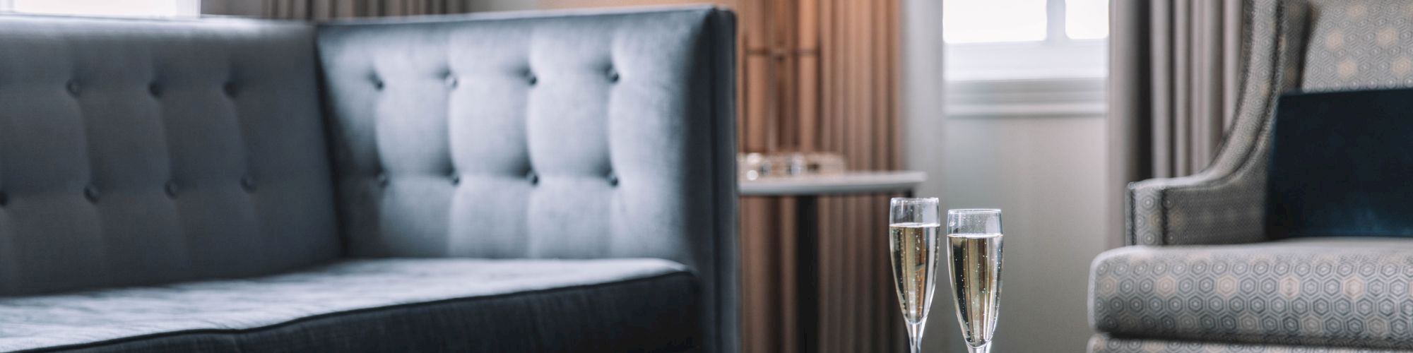 A cozy living room with a blue sofa, a round table with a book and two champagne glasses, next to a standing lamp and window curtains.