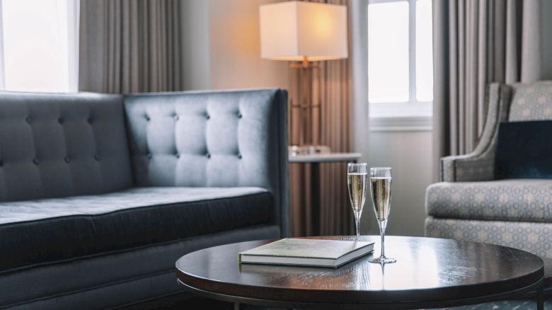 A cozy living room with a grey sofa, chairs, a round table with two champagne glasses, and a book, lit by a lamp near a window.