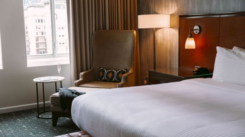 A cozy hotel room with a large bed, armchair, side table, and a window with city views. Warm lighting adds a comfortable ambiance.