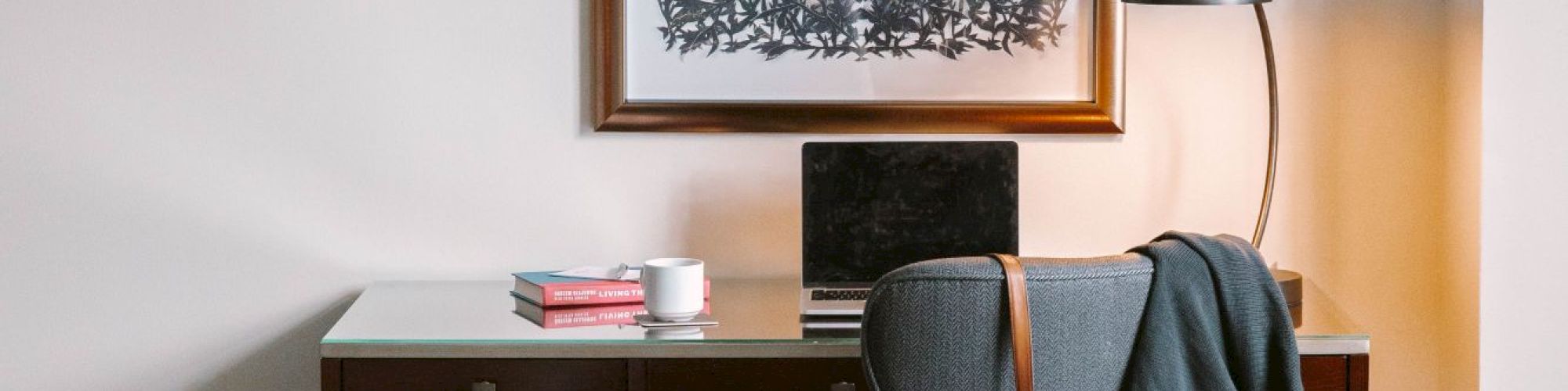 A cozy hotel room with a desk, chair, lamp, and artwork above, featuring a laptop, book, and a bag hanging on the chair.