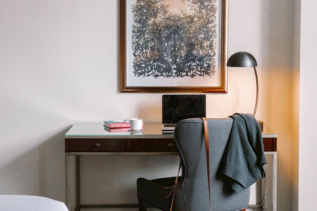 A cozy room with a desk, chair, lamp, and laptop; a jacket draped over the chair and bag hanging; artwork on the wall above.