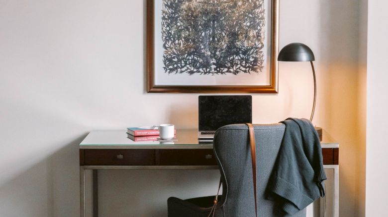 A cozy room with a desk, chair, lamp, and laptop; a jacket draped over the chair and bag hanging; artwork on the wall above.