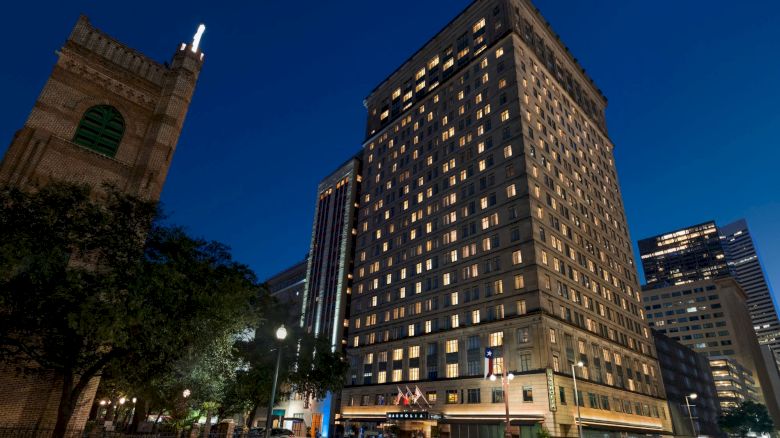 A tall, well-lit building at night with surrounding trees and other buildings. The scene includes a clear, dark blue sky.