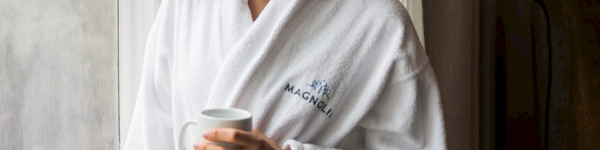 A person in a white bathrobe stands by a window holding a cup, looking outside.