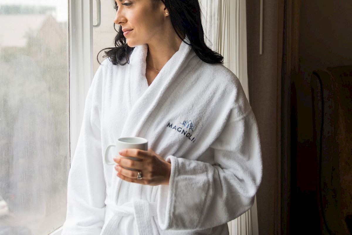 A woman in a white robe stands by a window, holding a cup, and looking outside, appearing thoughtful or relaxed.