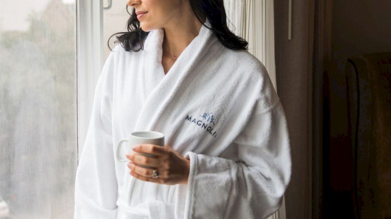 A woman in a white robe stands by a window, holding a cup, and looking outside, appearing thoughtful or relaxed.