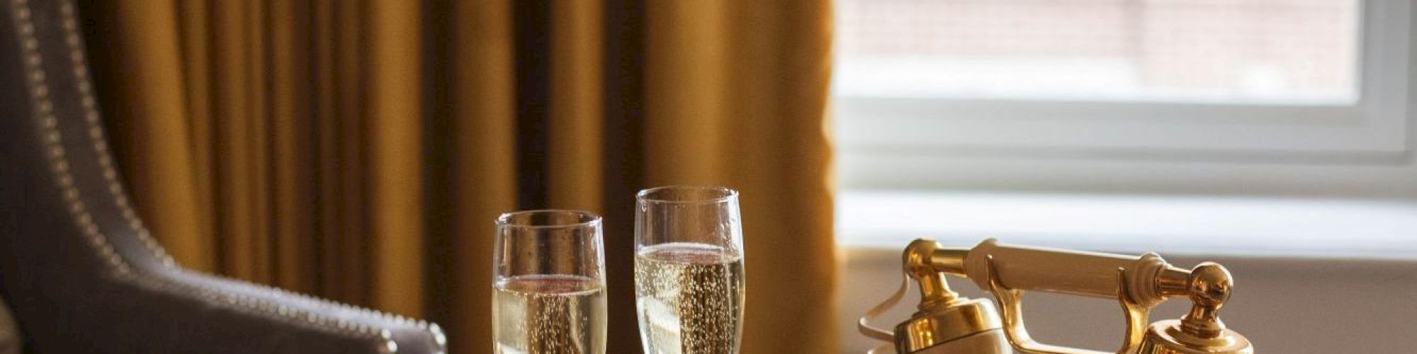 Two champagne glasses and a vintage-style telephone sit on a tray on a bed near a window with yellow curtains.
