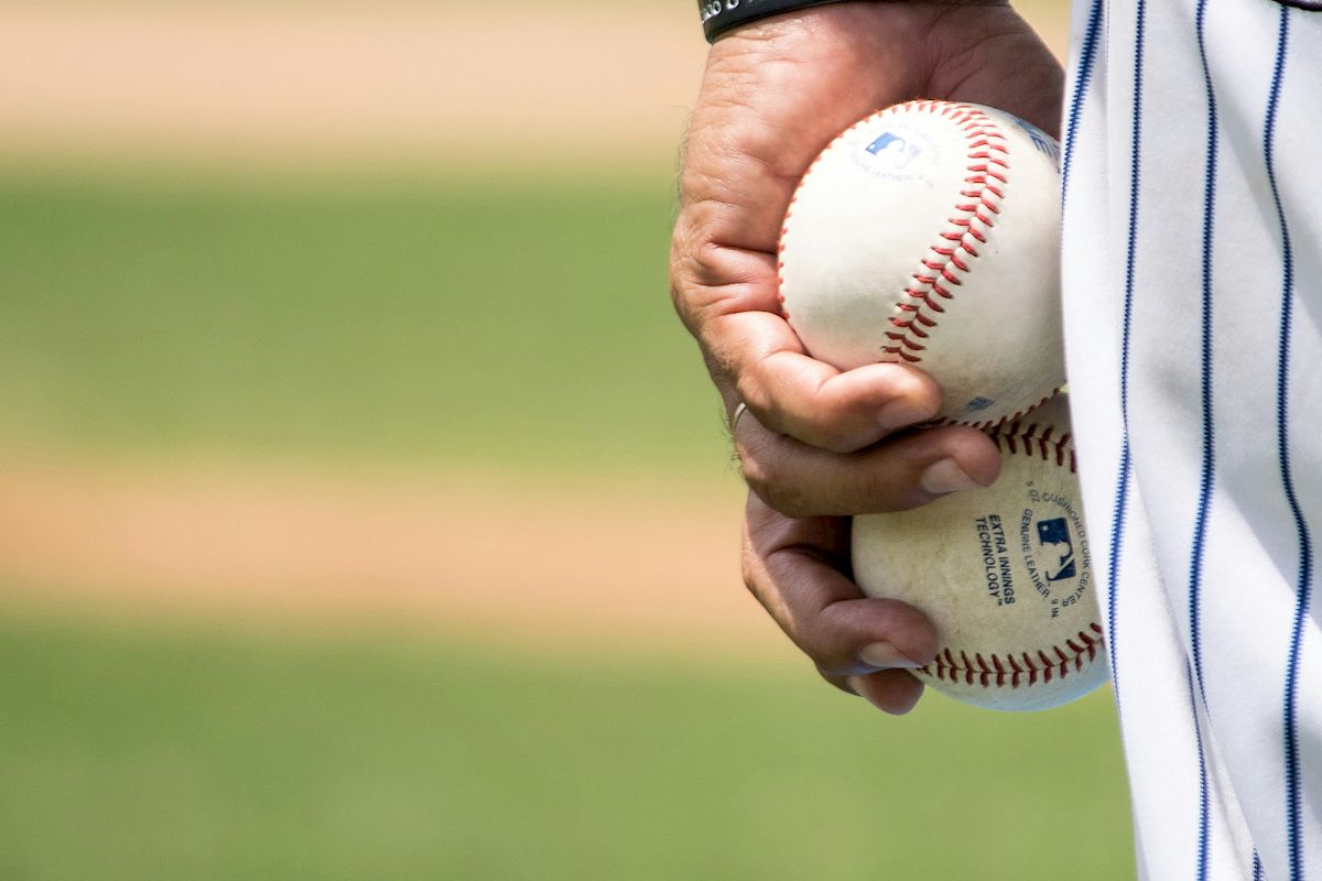 A person wearing a striped jersey holding two baseballs with a baseball field in the background.