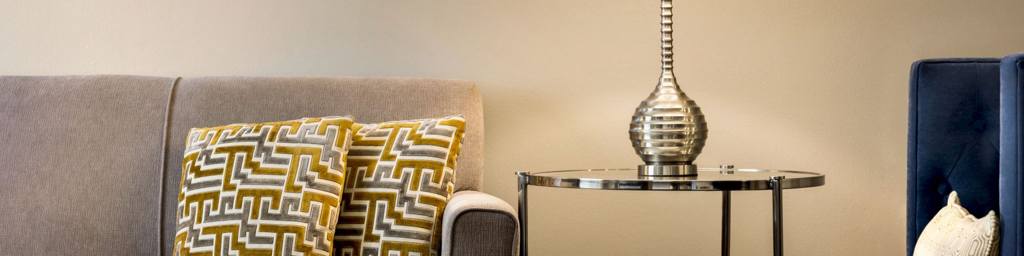 A cozy living room with a sofa, patterned cushions, a coffee table, a lamp on a side table, books, and a chair with a cushion.