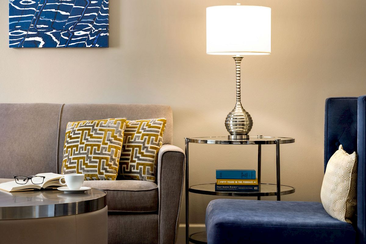 A cozy living room with a sofa, patterned cushions, a table lamp on a side table, and a chair. A book and cup are on the coffee table.