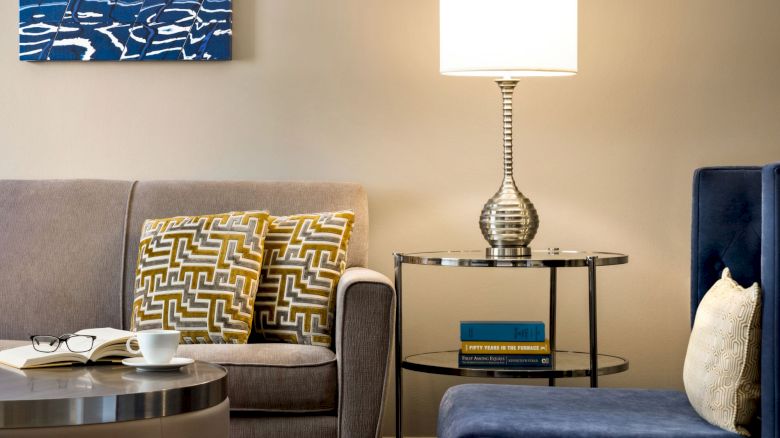 A cozy living room with a sofa, patterned cushions, a table lamp on a side table, and a chair. A book and cup are on the coffee table.