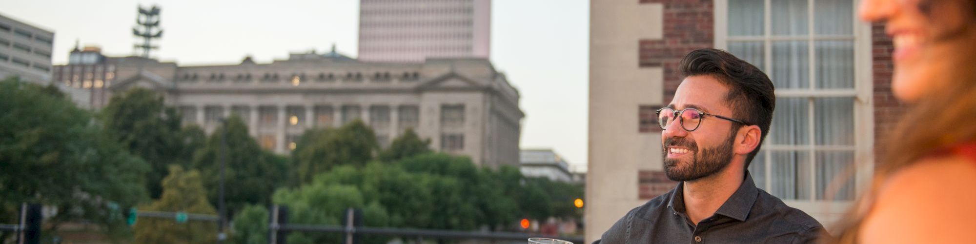 Two people enjoying drinks on a balcony with a cityscape in the background.