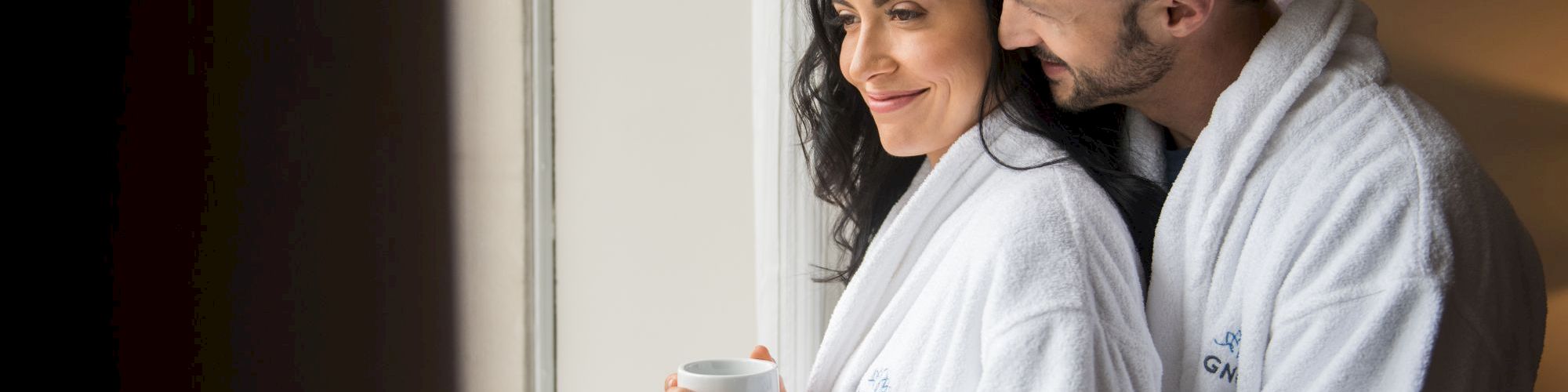 A couple in white robes stands by a window, with one holding a mug, sharing a tender moment in natural light.