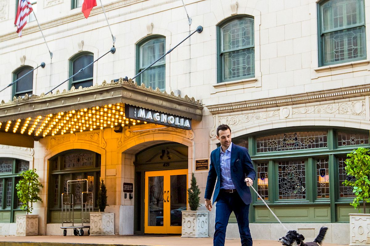 A man in a suit walks a black and white dog across the street outside a hotel building with flags and an awning.