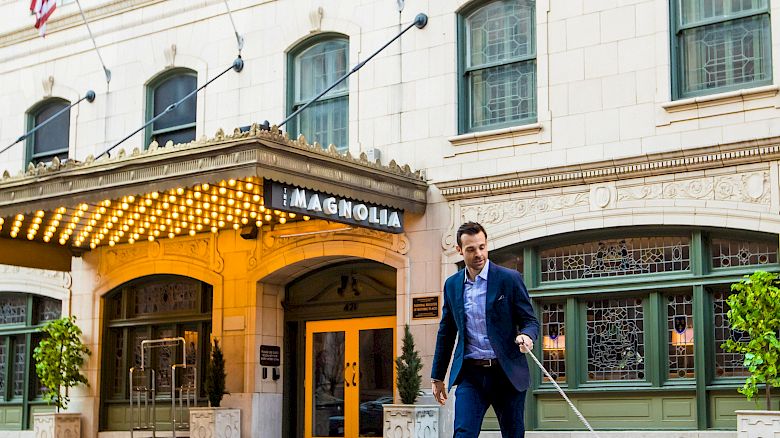 A man in a suit walks a black and white dog across the street outside a hotel building with flags and an awning.