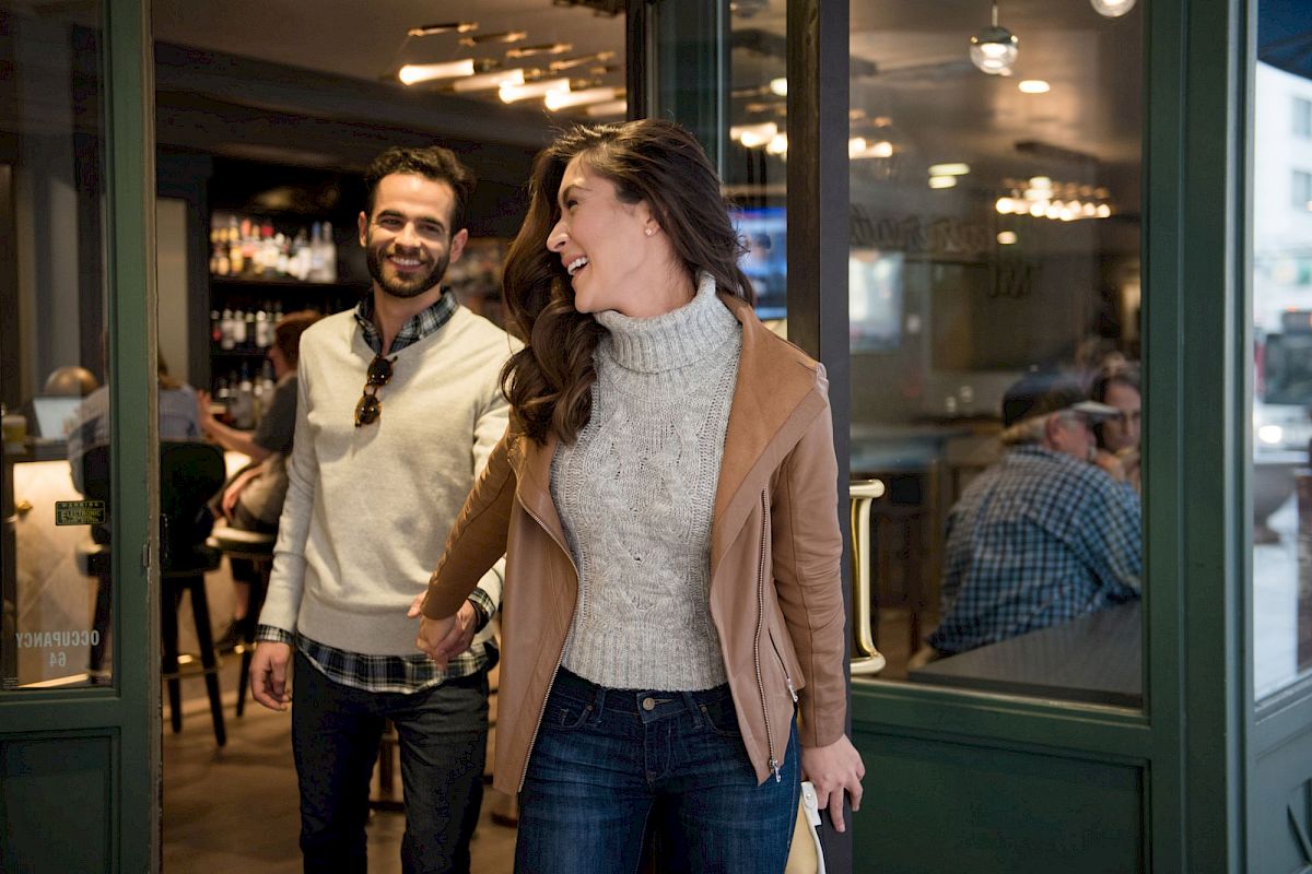 A woman and a man are smiling while leaving a cafe. She holds the door open as they walk out, appearing cheerful.