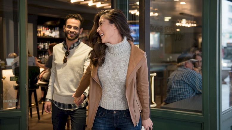 A woman and a man are smiling while leaving a cafe. She holds the door open as they walk out, appearing cheerful.