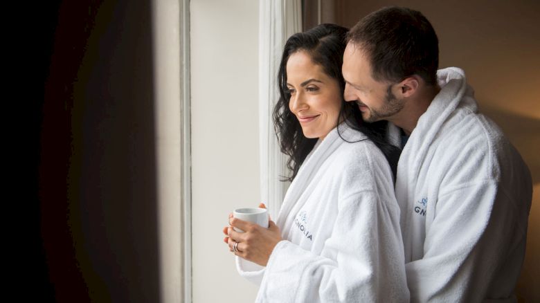 A couple in white robes is standing by a window; one holds a mug, both appear relaxed and content in a cozy setting.