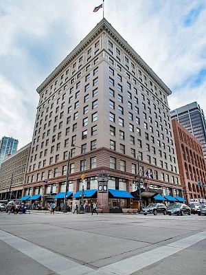The image shows a tall brick building with a flag on top at a city intersection with blue awnings and smaller buildings nearby.