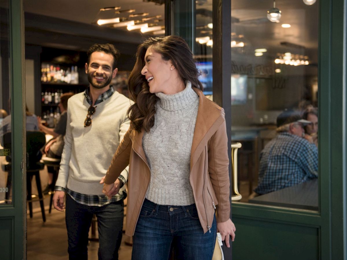 A couple is joyfully exiting a restaurant, holding hands, with people visible inside and modern lighting in the background.