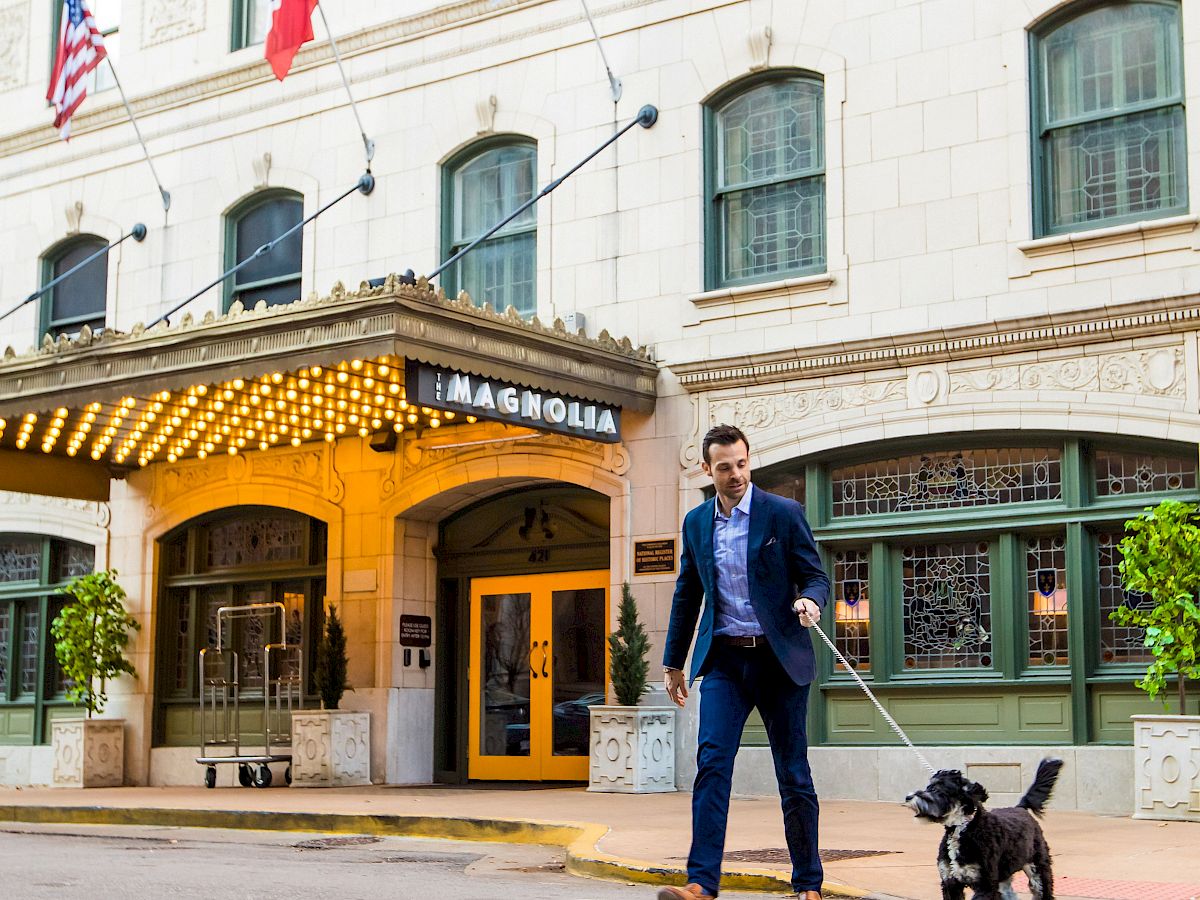 A person in a suit walks a dog across a street in front of a building with flags and a decorative awning.