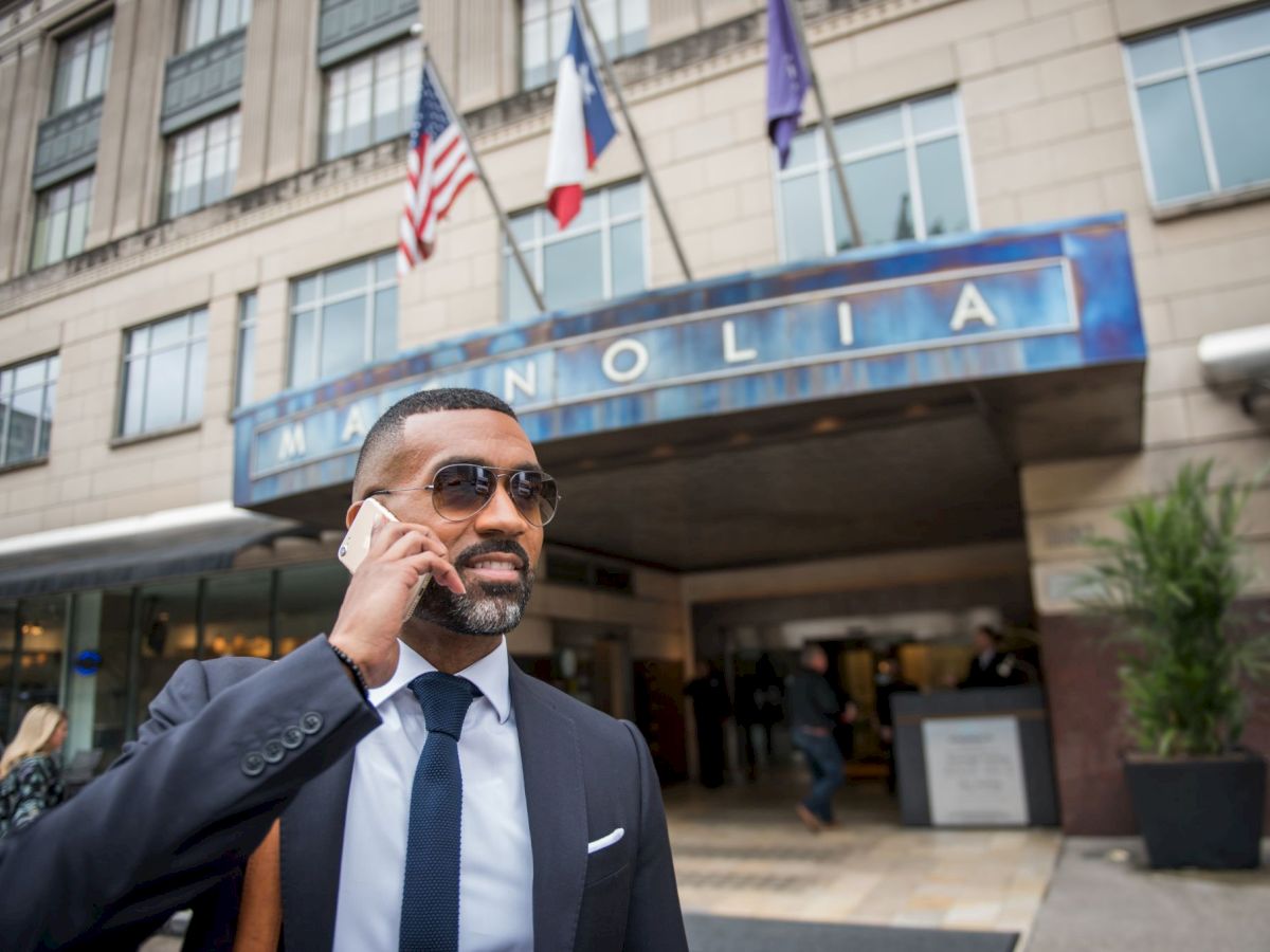 A man in a suit is talking on a phone in front of a building with flags and a sign saying 