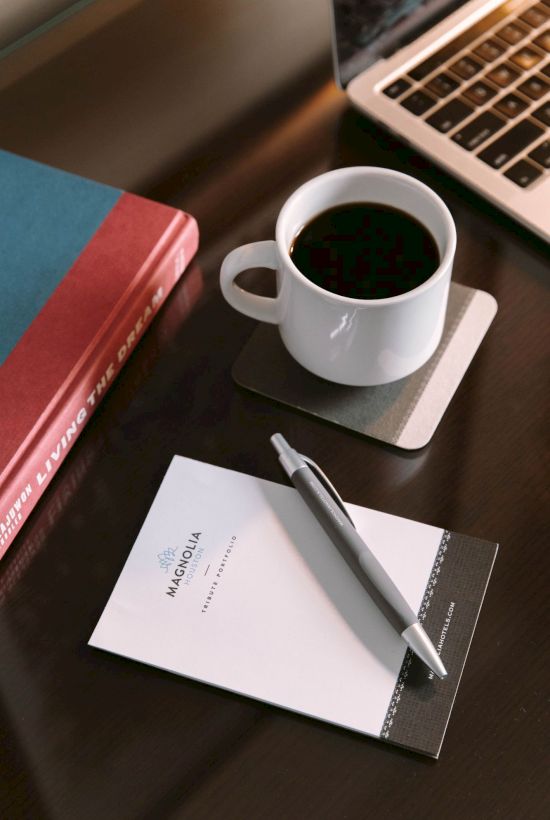 A notebook, pen, coffee cup on a coaster, book, and laptop on a dark desk.