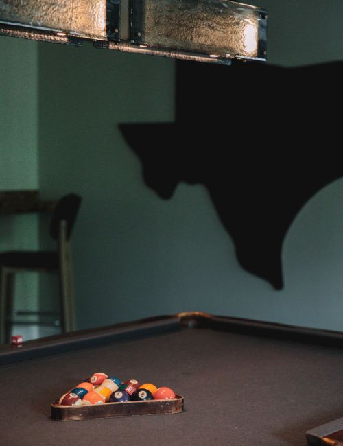 A billiards table with racked balls sits under a light fixture, with a Texas-shaped wall decoration in the background.