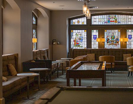 A cozy lounge with stained glass windows, sofas, tables, and a shuffleboard table in a warmly lit setting.