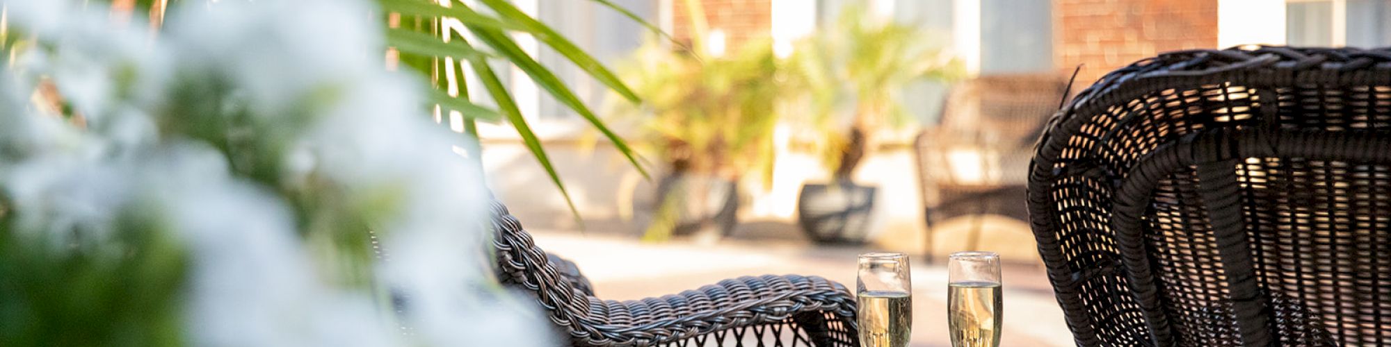 Outdoor seating area with wicker chairs, a small table holding two champagne glasses, surrounded by greenery and flowers in soft focus.