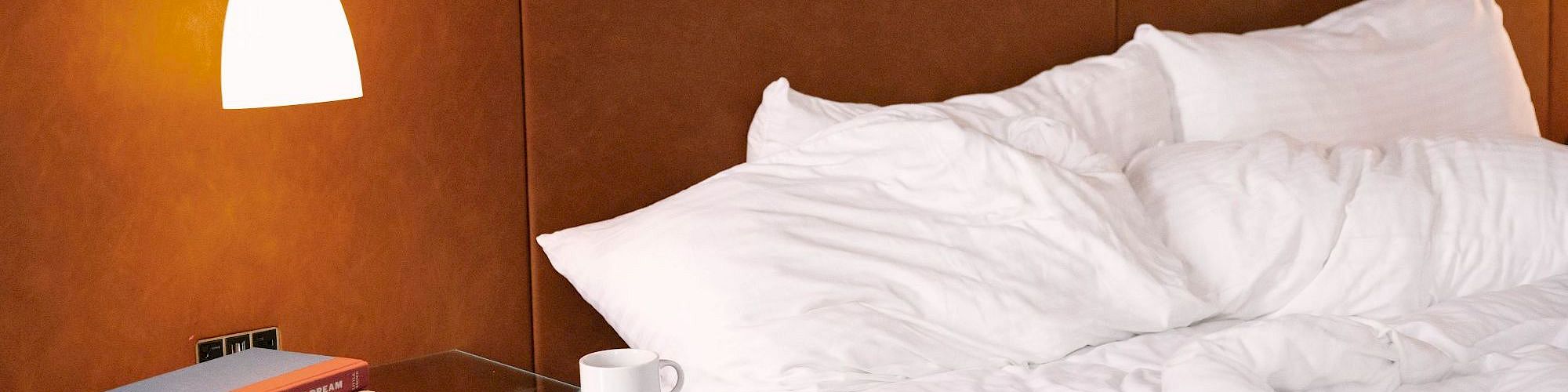 A cozy bedroom with a neatly made bed, wall lamp, books, and a cup on the nightstand, under soft, ambient lighting.