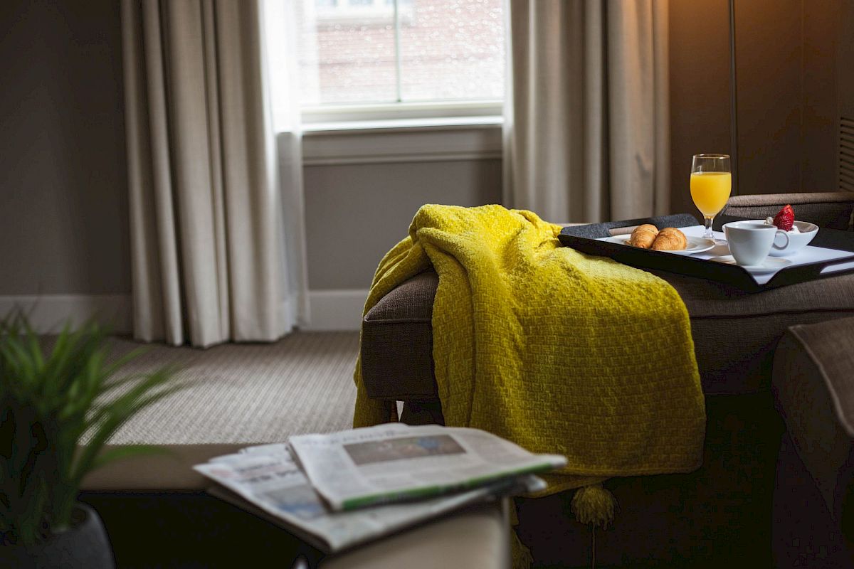 A cozy room with a yellow blanket, a tray with breakfast items, and newspapers on a chair, next to a window with curtains.