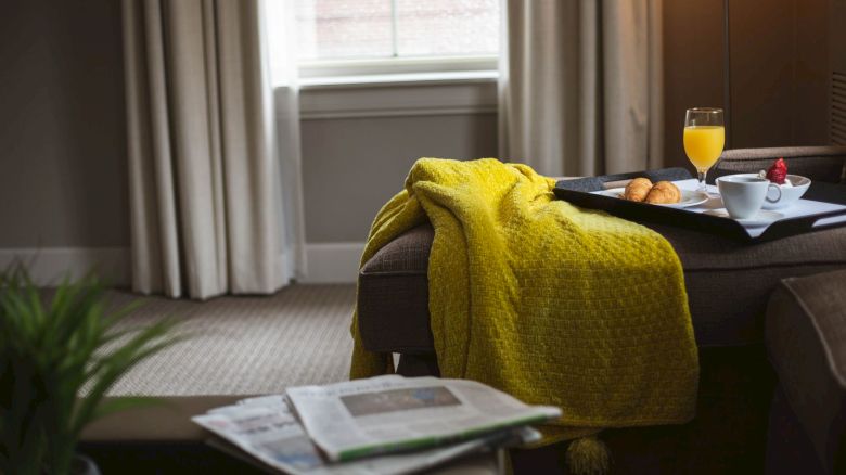 A cozy room with a yellow blanket, a tray with breakfast items, and newspapers on a chair, next to a window with curtains.