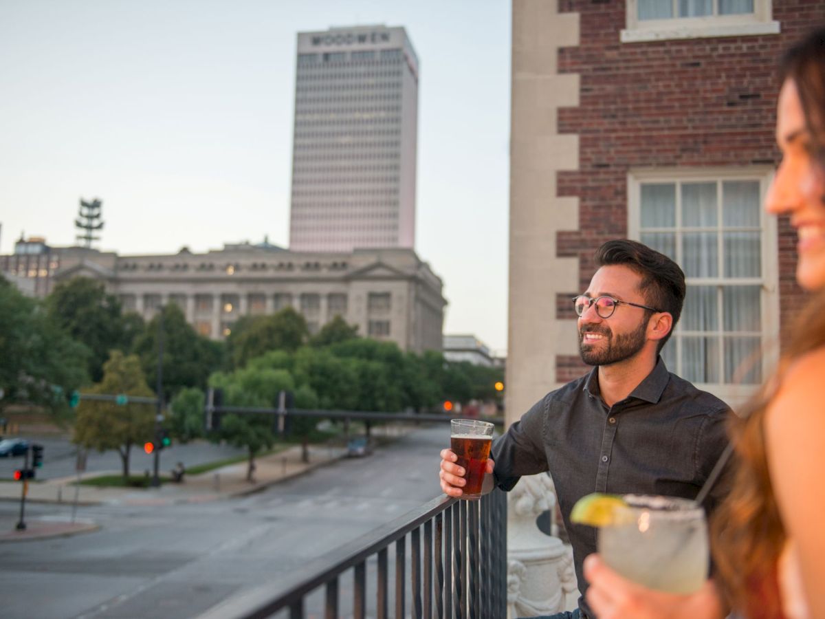 Two people are enjoying drinks on a balcony overlooking a city street with a tall building in the background.