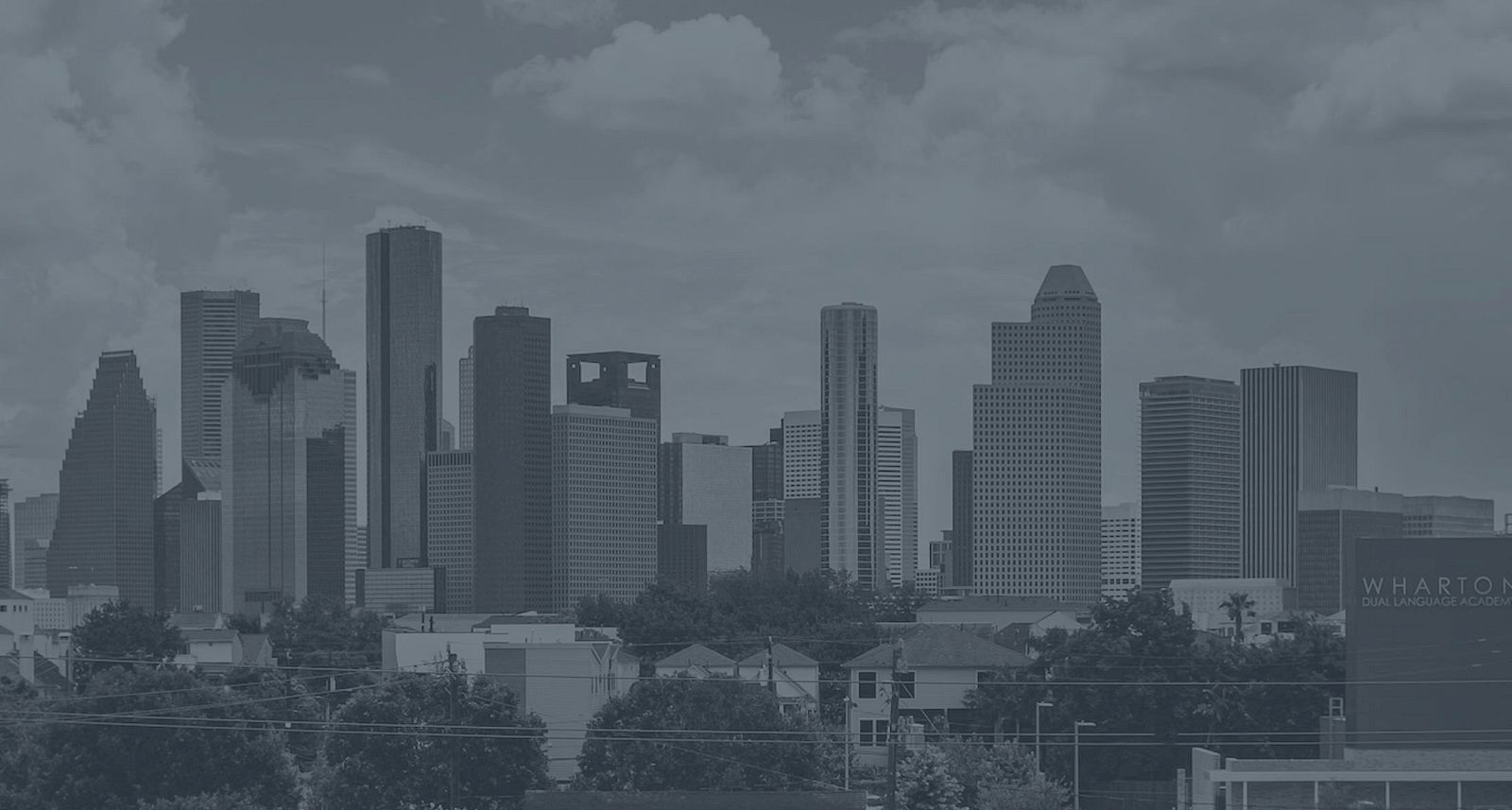 A city skyline with numerous tall buildings against a cloudy sky, featuring trees and smaller structures in the foreground.