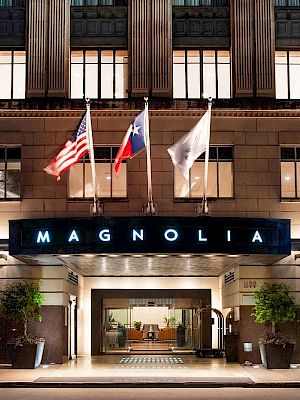 The image shows the entrance of a Magnolia building with three flags above. The facade is illuminated, and large windows are visible.
