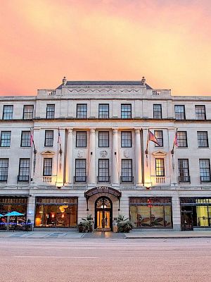 A large, elegant building with multiple windows and flags, set against a vibrant sunset sky. There are cars parked in front.
