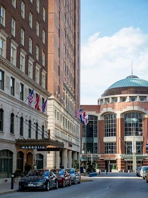 A city street view with brick buildings, flags, and parked cars. The architecture includes domed and classic styles under a blue sky.