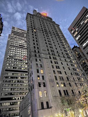 A cityscape at dusk features tall buildings with lights, street-level shops, and trees adorned with decorative lights.